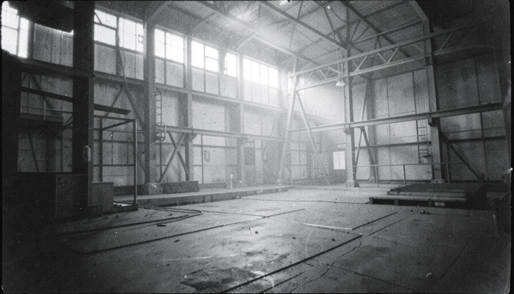 Fotografía analógica en blanco y negro con grano visible de una nave industrial vacía, estructuras metálicas y luz difusa entrando por ventanas altas, ambiente sobrio y silencioso