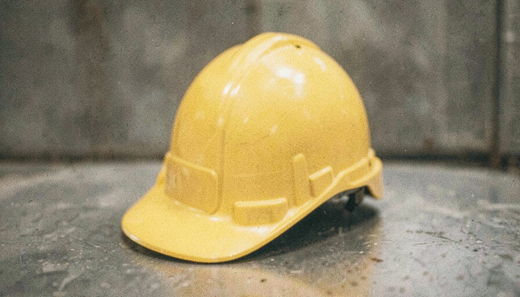 Fotografía analógica granulada de un casco de seguridad industrial de color amarillo apagado sobre una superficie metálica gris, con luz tenue y tonos tierra que transmiten seriedad y cuidado