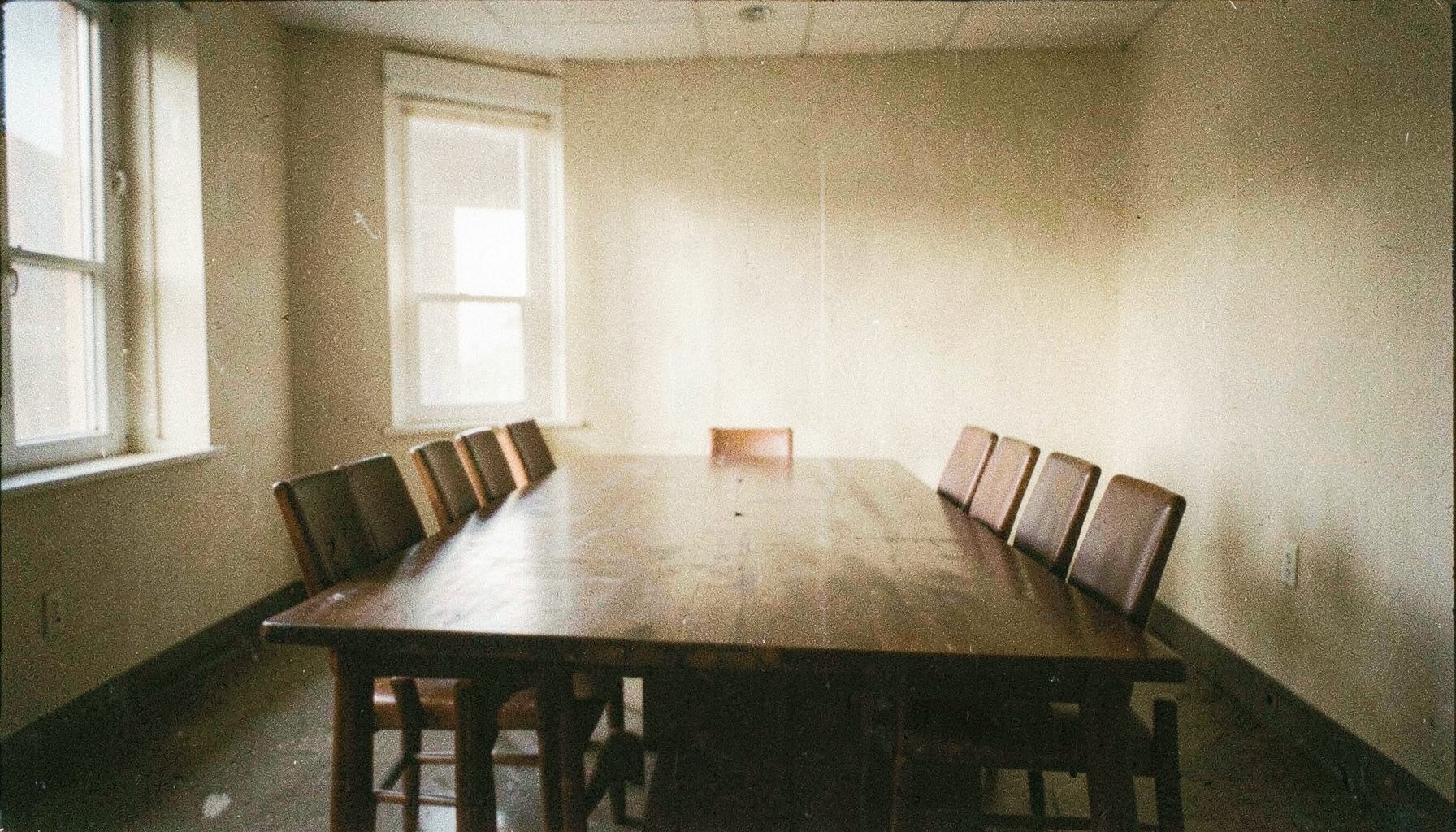 Fotografía analógica granulada de una sala de reuniones austera con una mesa larga de madera oscura, sillas alineadas y luz suave entrando por una ventana, en tonos marrones cálidos y beige que transmiten seriedad y calma